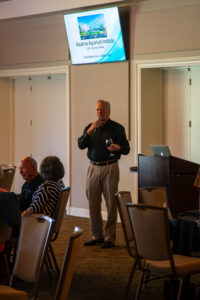 Man speaking into a microphone under a video screen